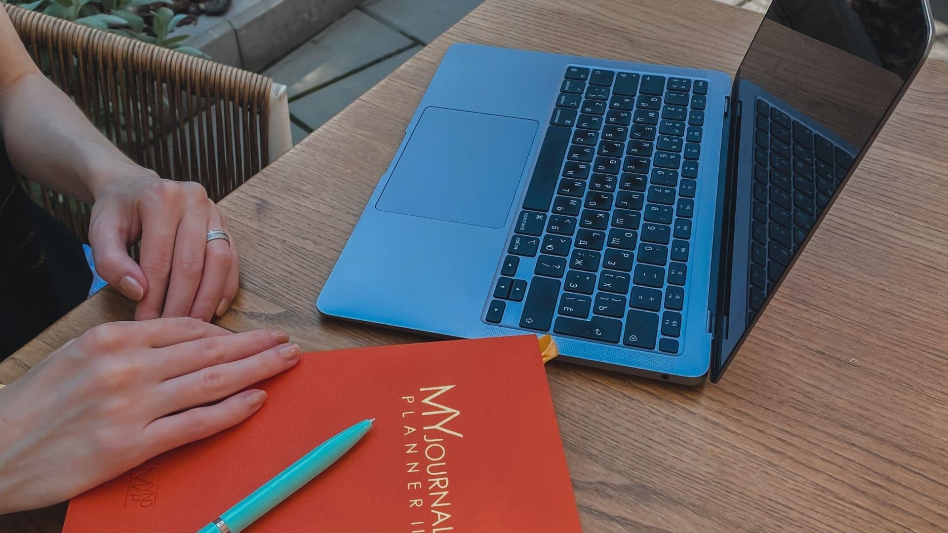 Tetyana is sitting at a table outdoors. Her hands are on an orange planner, which is called MyndMap. There is a Tiffany pen on her planner. There is a laptop near her planner.