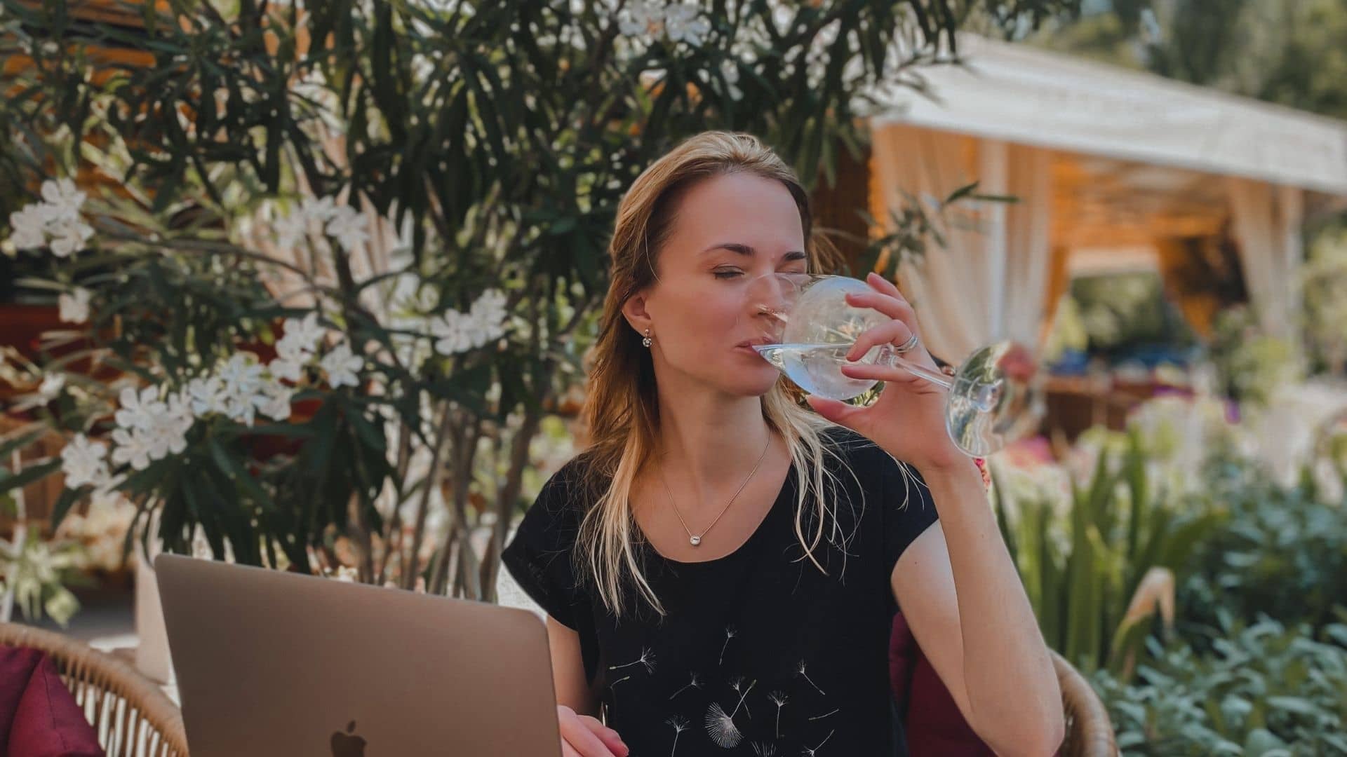 Tetyana is drinking water from a wine glass with her eyes closed. She is sitting at a table with a laptop. There are green trees on the background. 